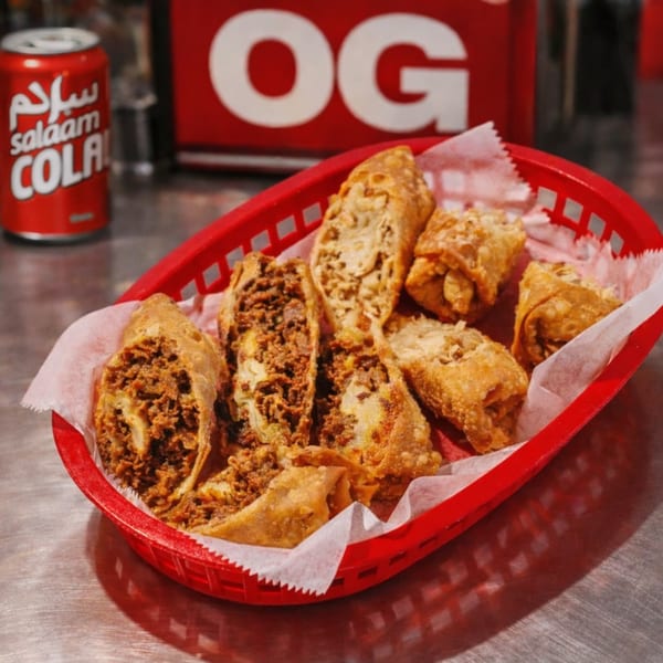 A red basket of crispy egg rolls, some cut open to show a savory filling, with a Salaam Cola can.