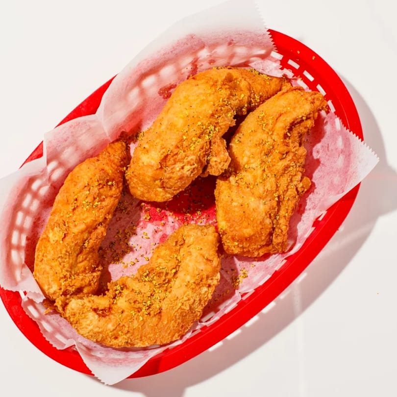 Four crispy fried chicken tenders, seasoned with yellow spices, in a red plastic basket with white paper lining.